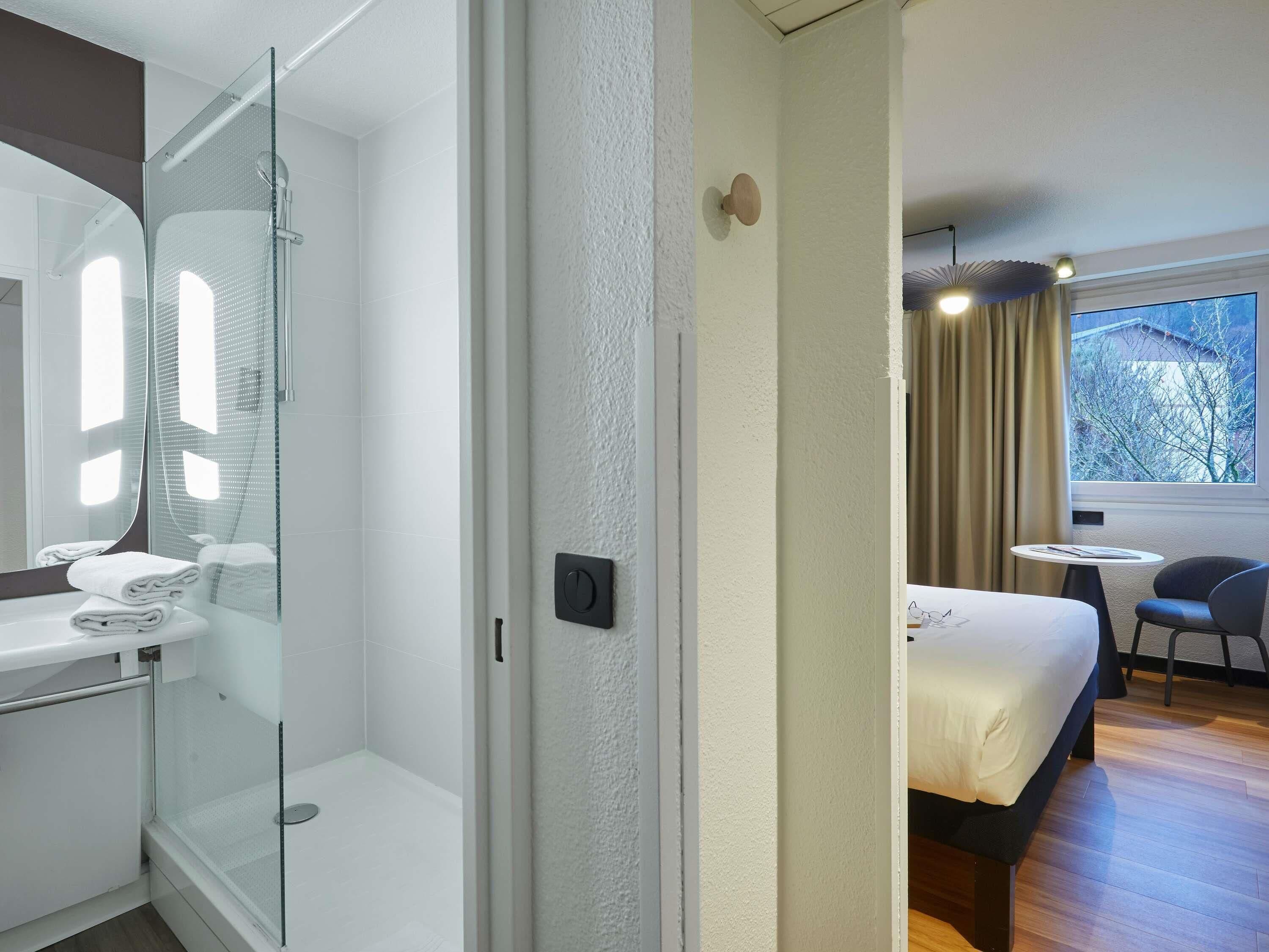 A hotel room with a glass-enclosed shower, a bed with a white duvet, a small round table, and a chair.