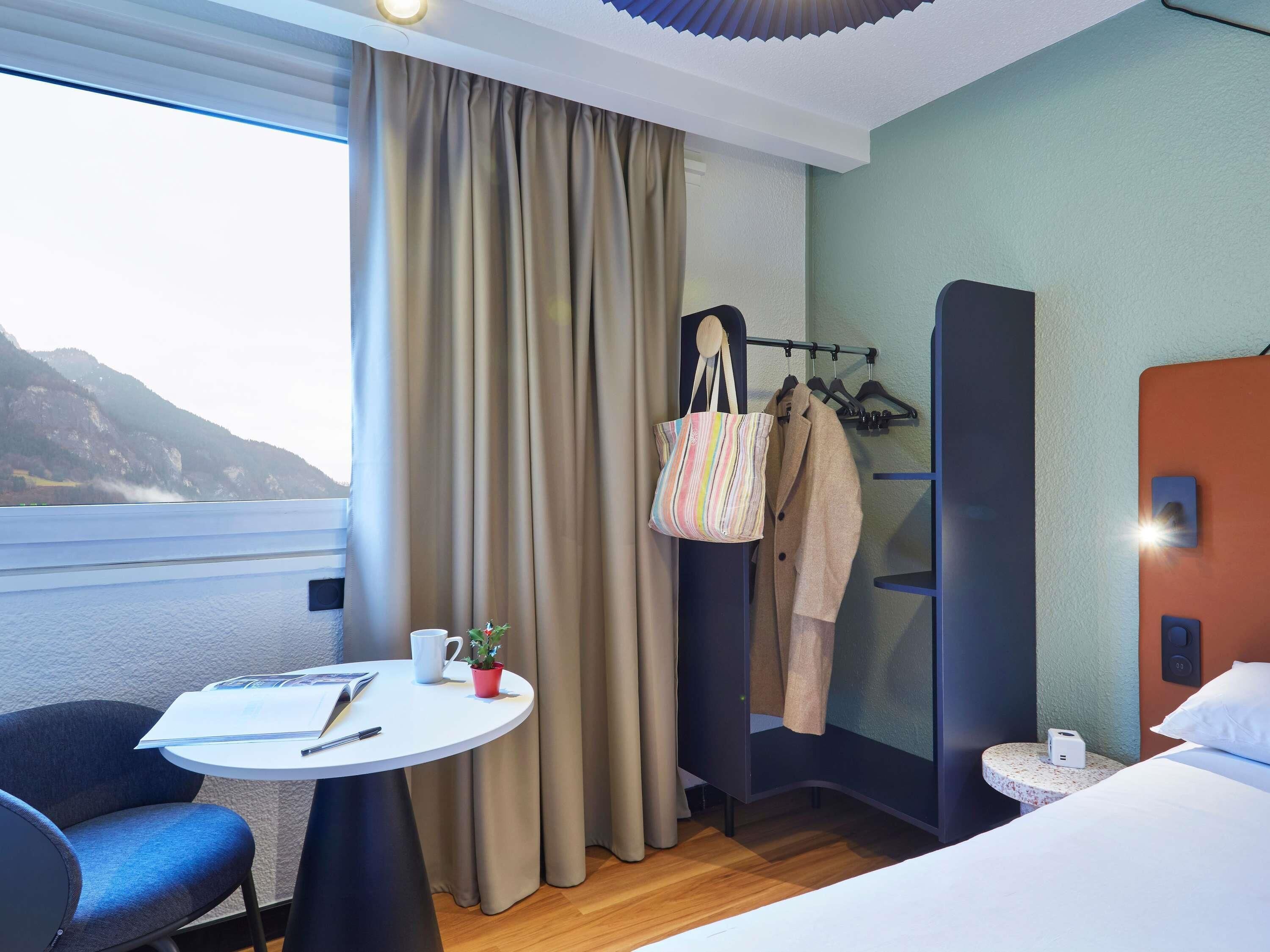 Una habitación de hotel con una cama, una mesita con una silla, un perchero con abrigo y bolso, y una ventana con cortinas.