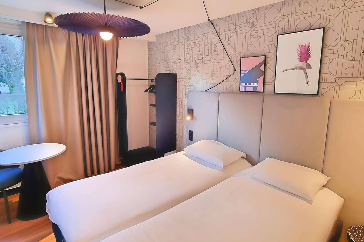 A hotel room with a bed, a desk, a chair, a standing lamp, and a wall with geometric patterns.
