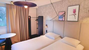 A hotel room with a bed, a desk, a chair, a standing lamp, and a wall with geometric patterns.
