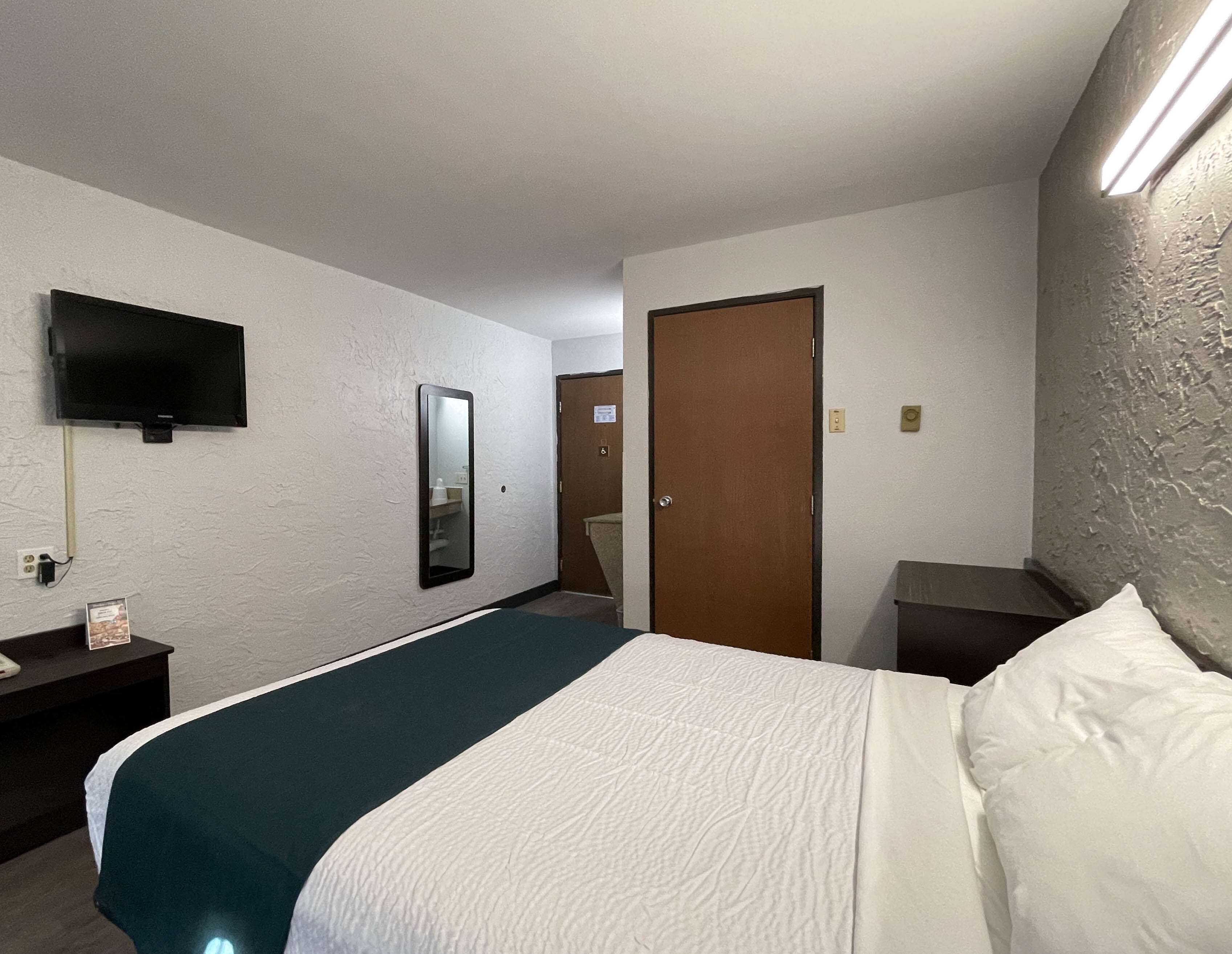 Une chambre d’hôtel avec un lit, un téléviseur fixé au mur, un miroir, une porte et une table de chevet avec une lampe.