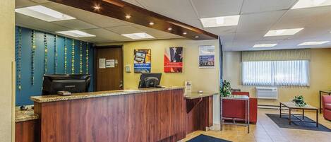A hotel reception area with a wooden reception desk, a computer, and a seating area with a red chair and a small table.