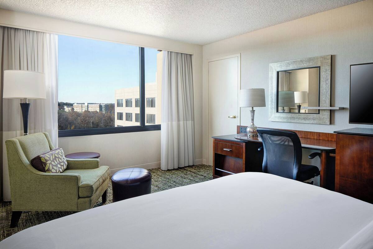 A hotel room with a large bed, a desk with a chair, a small table, a lamp, and a TV.