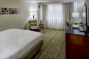 A hotel room with a large bed, a desk with a computer, a chair, a small table with a lamp, and a window with curtains.