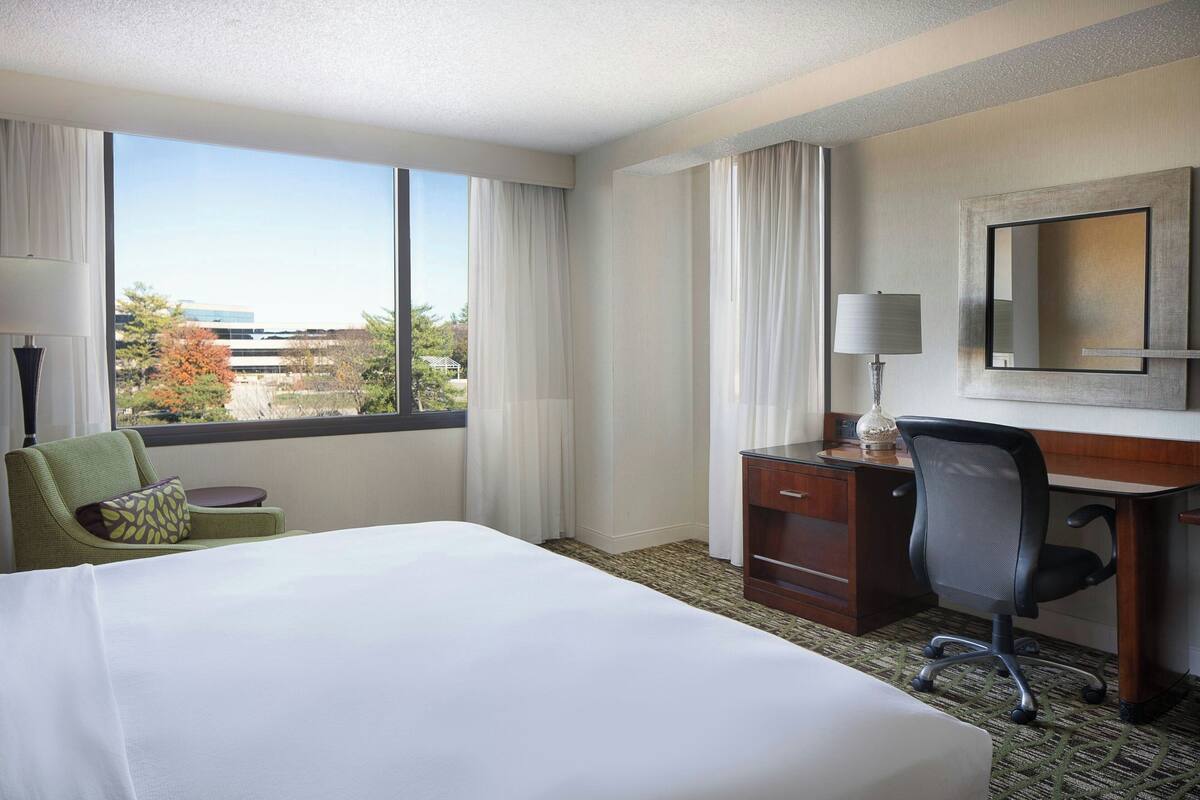A hotel room with a large bed, a desk with a chair, a lamp, and a view of the city.