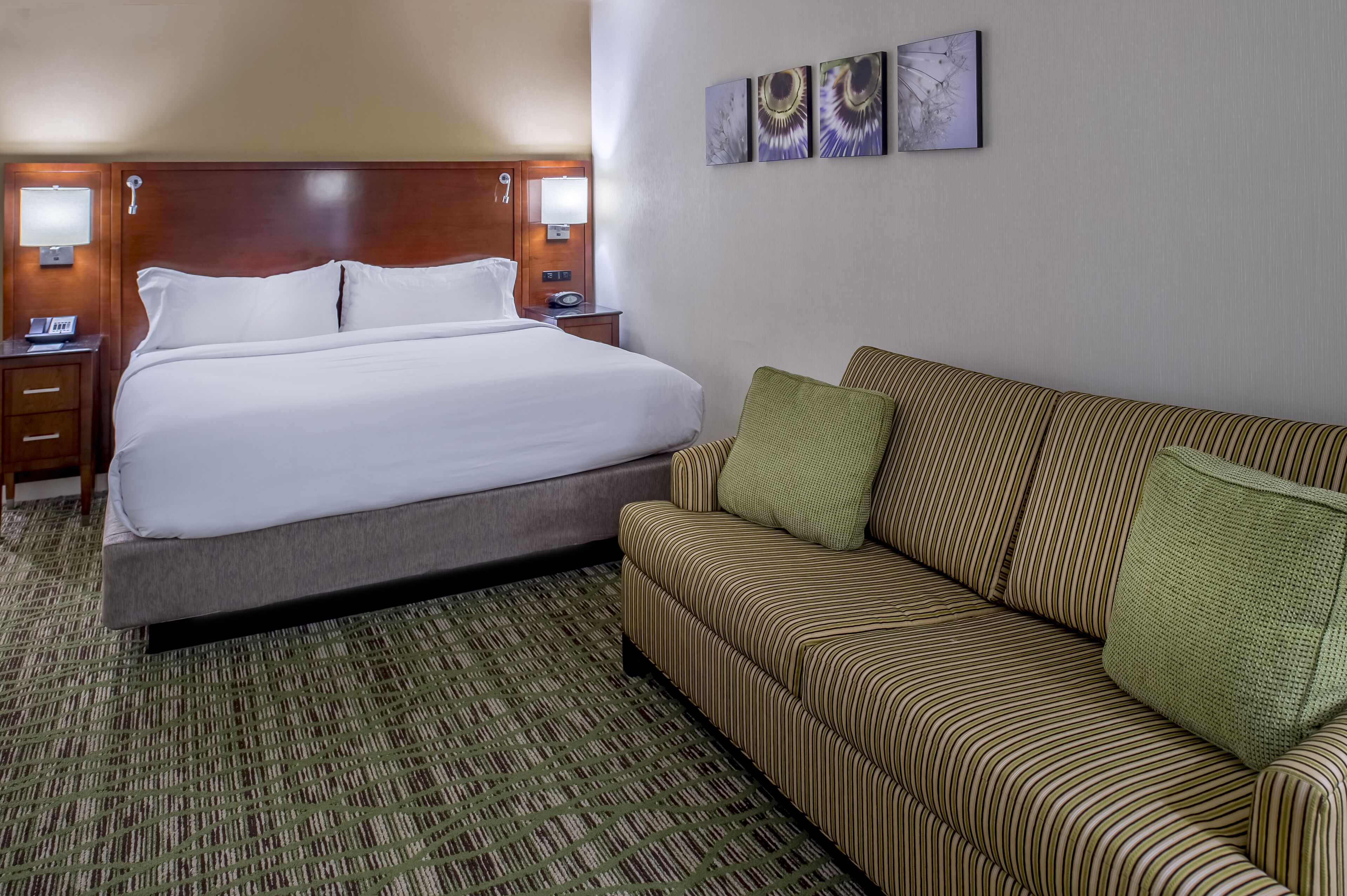 A hotel room with a large bed, a striped sofa, a wooden headboard, two bedside tables with lamps, and three wall-mounted artworks.