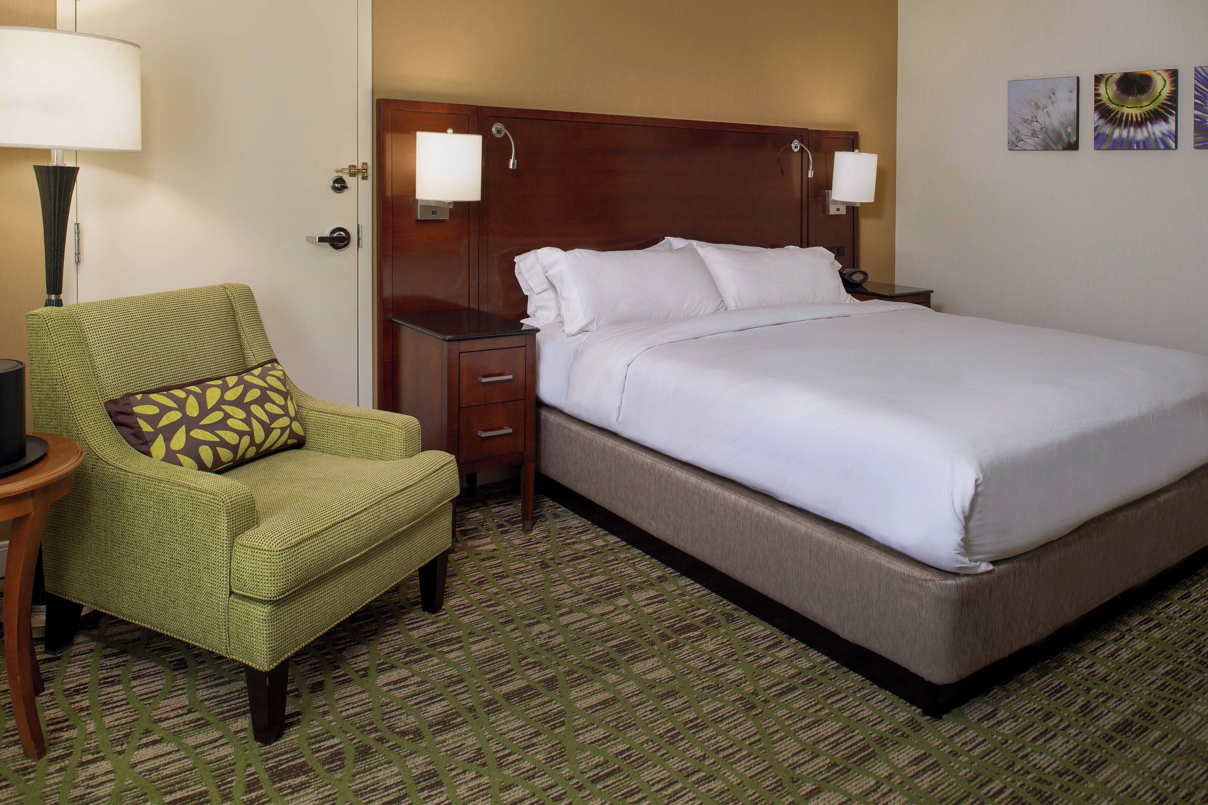 A hotel room with a large bed, a green armchair, a nightstand, and a lamp.