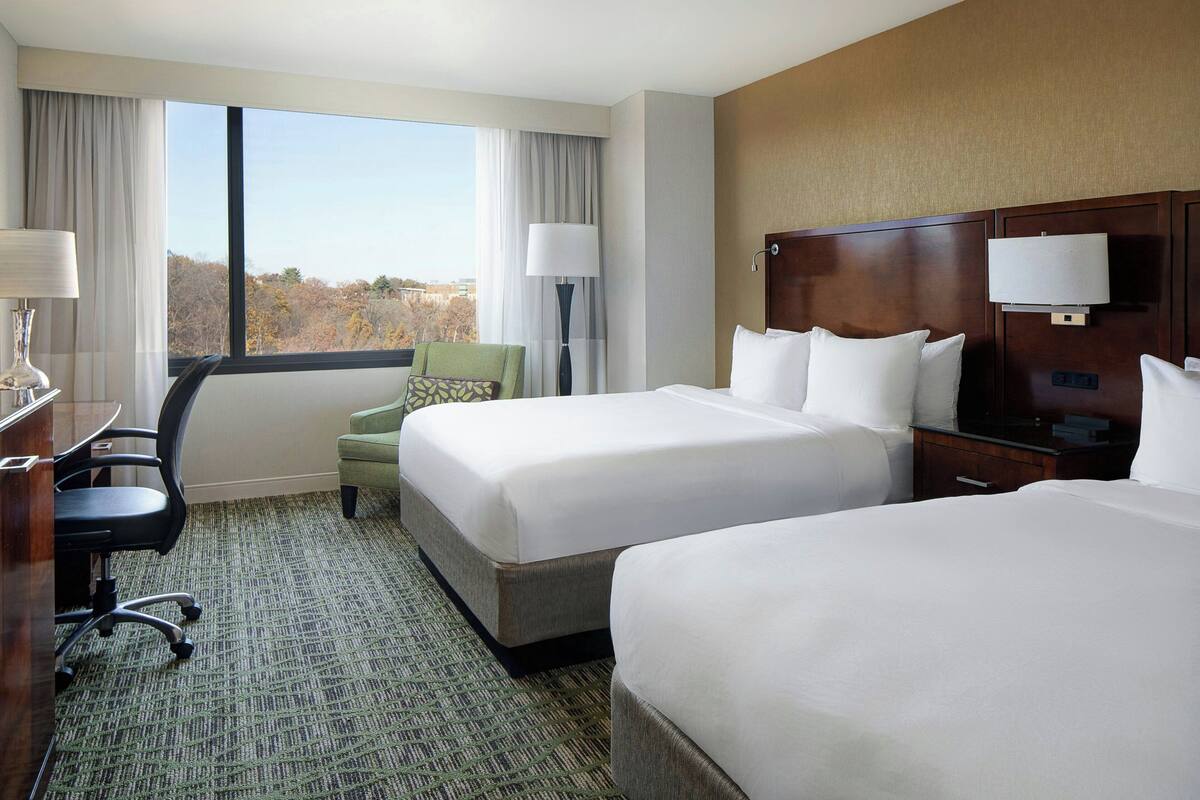 A hotel room with two beds, a desk, a chair, and a view of the outdoors.