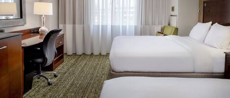 A hotel room with two beds, a desk with a chair, a lamp, and a large window with curtains.