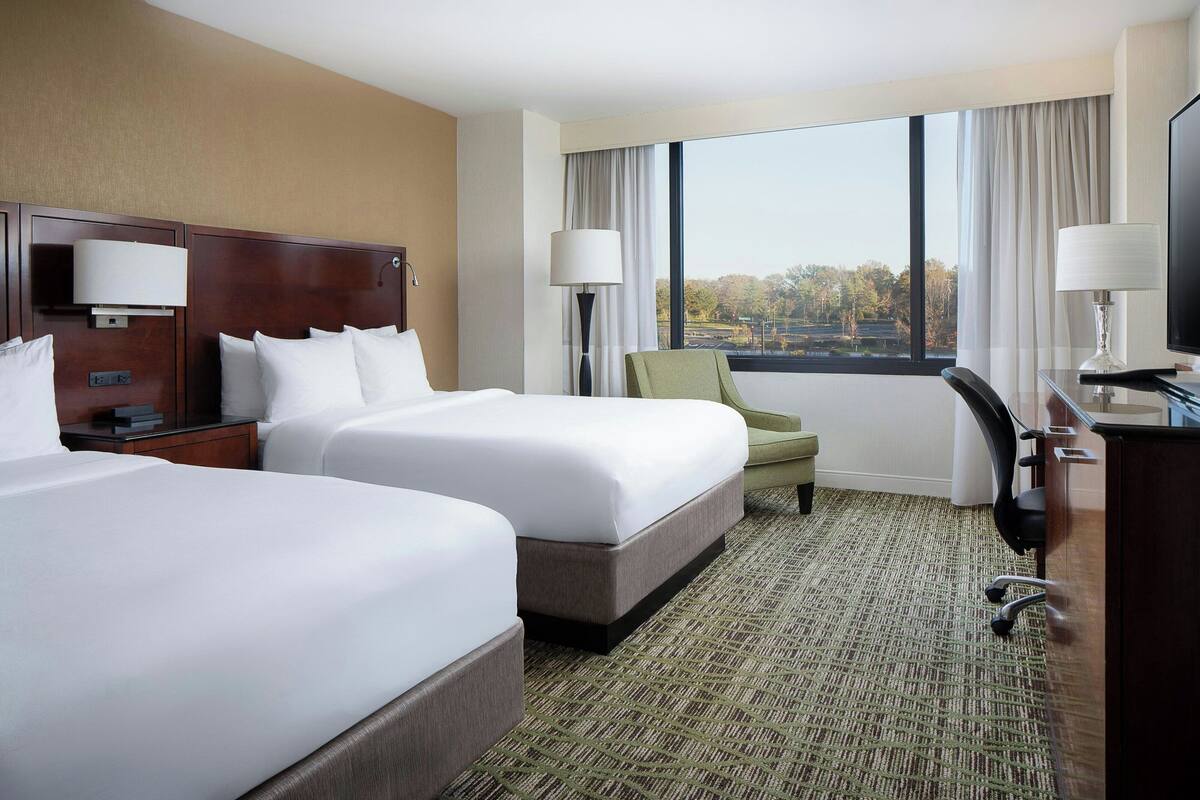 A hotel room with two beds, a desk, a chair, and a large window offering a view.