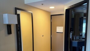 Une chambre d’hôtel comprenant un lit, un banc, un miroir, une salle de bain avec un lavabo et un miroir, et une porte donnant sur une autre pièce.