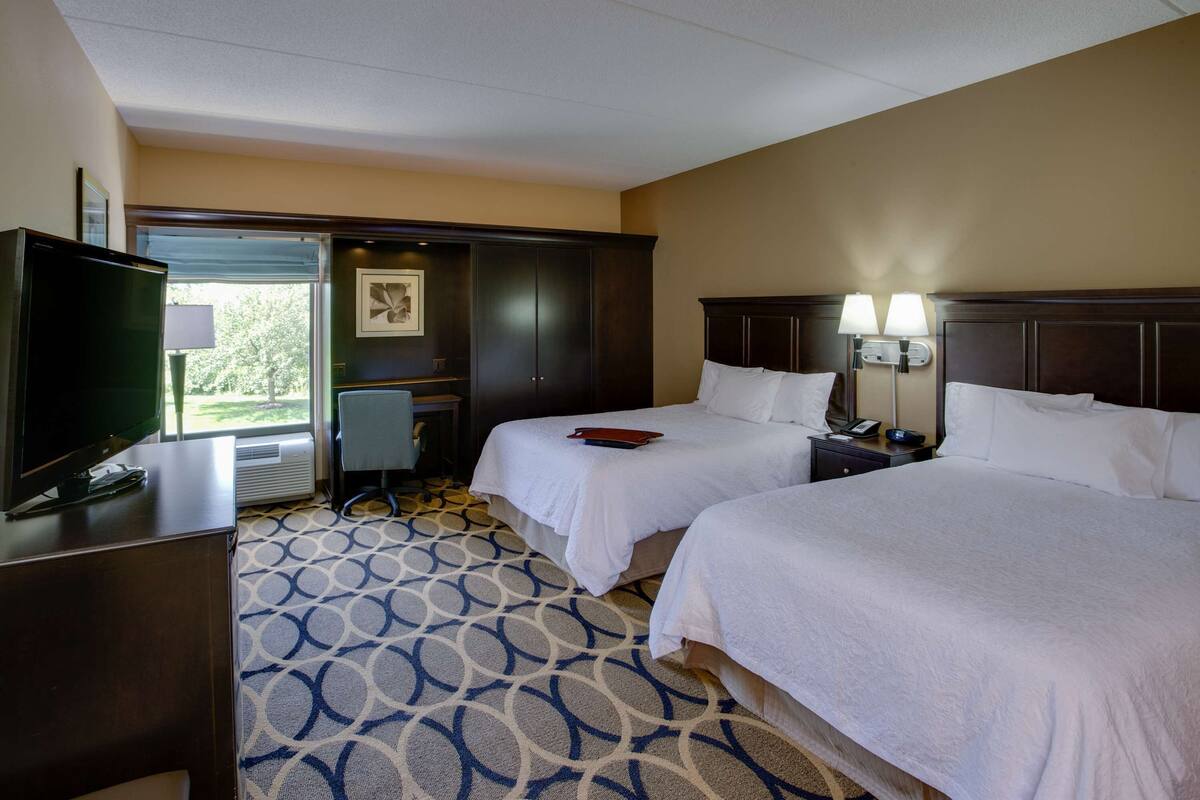 A hotel room with two beds, a television, a desk, and a window with a view.