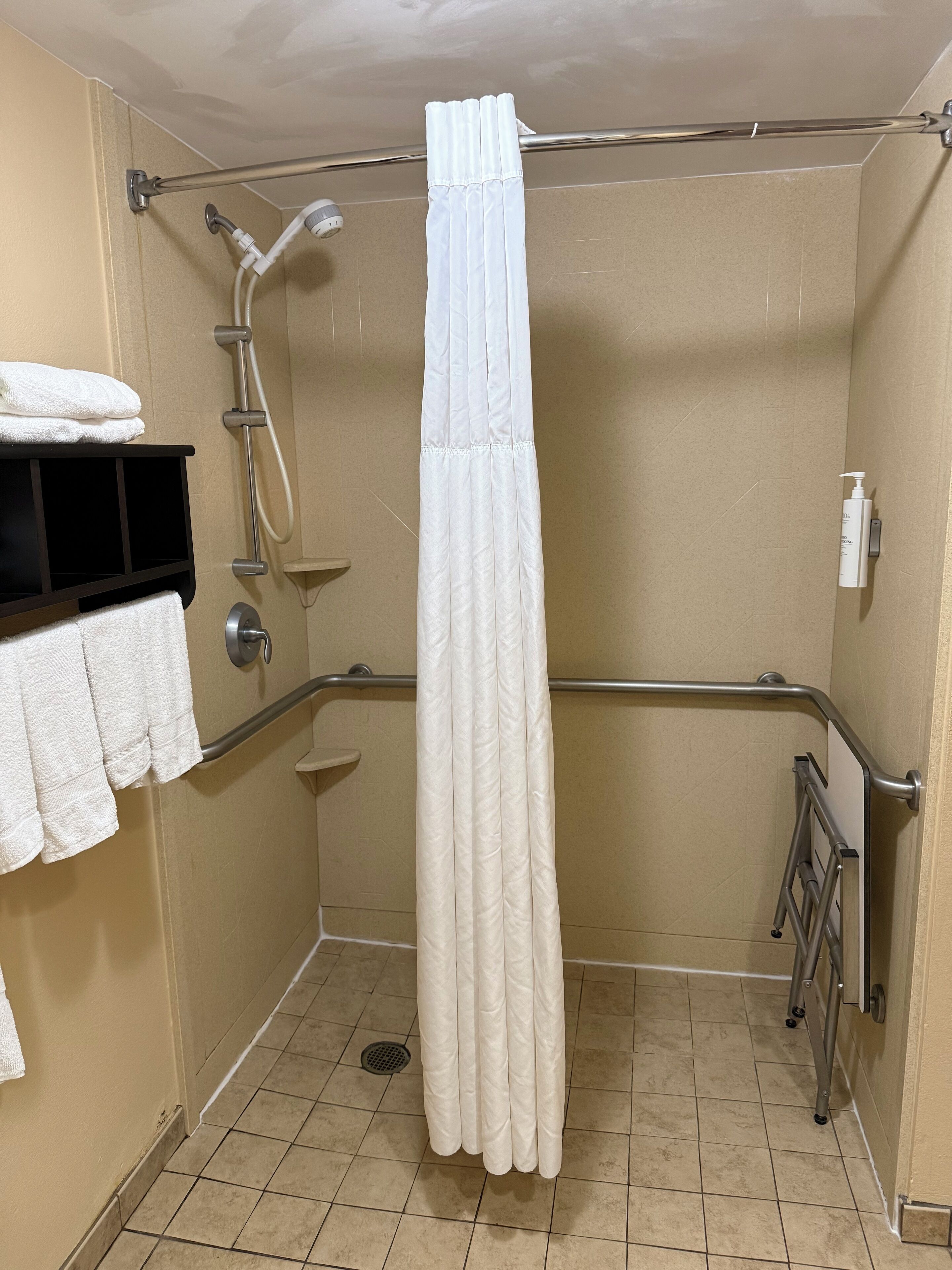 Un baño con cortina de ducha blanca, barra de apoyo, toallero y ducha de mano.