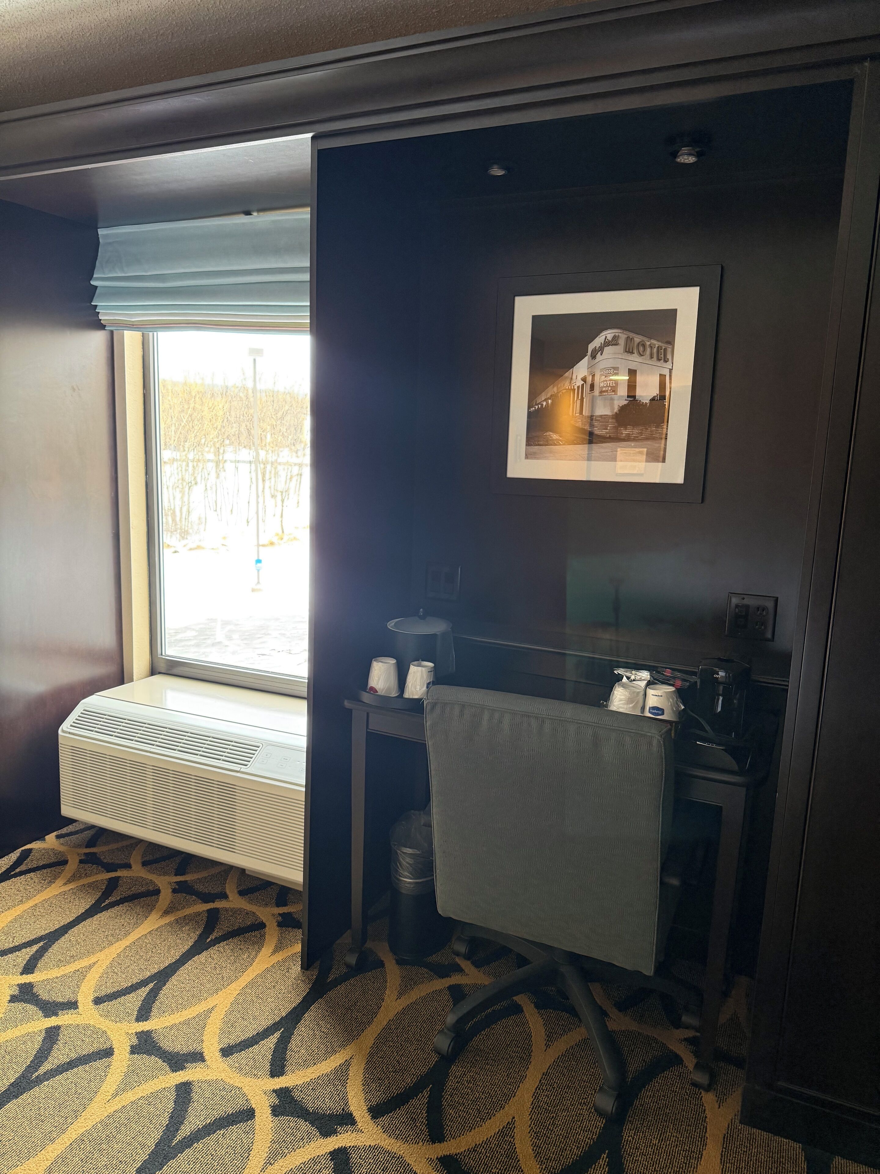 Una habitación de hotel con ventana, un radiador, una pequeña mesa con lámpara, una silla y un cuadro en la pared.