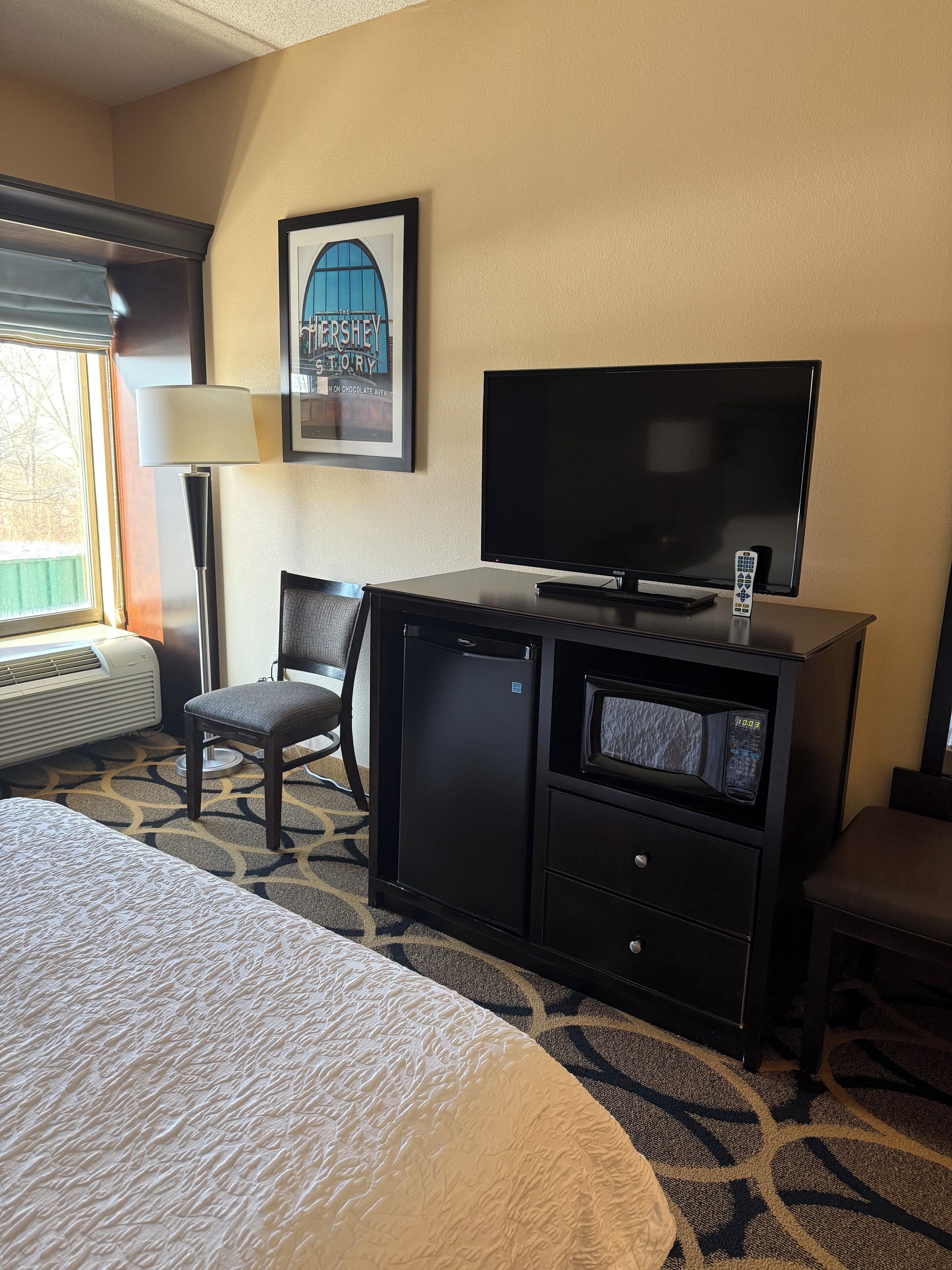 A hotel room with a bed, a desk with a television, a chair, a framed picture on the wall, and a window with blinds.