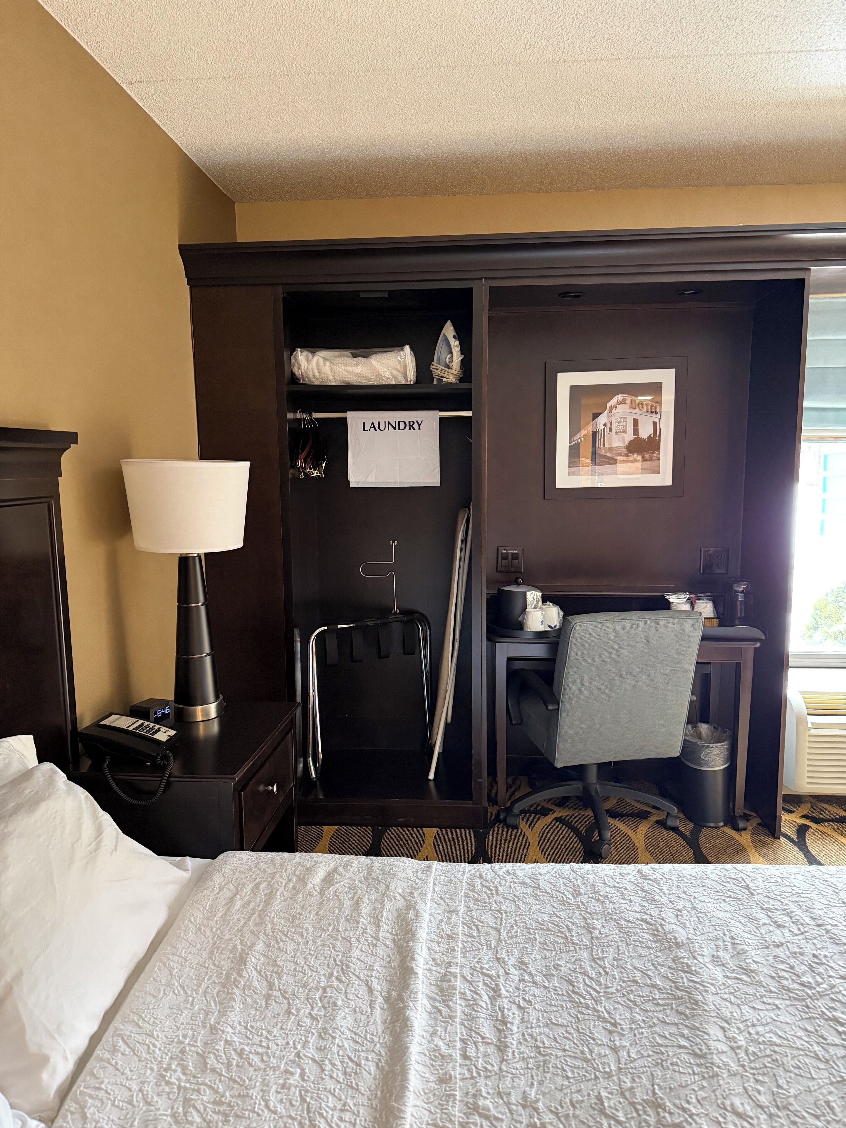 A hotel room with a bed, a desk with a chair, a lamp, a coat rack, a trash can, and a laundry basket.