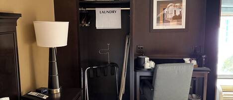 Een hotelkamer met een bed, een bureau met een stoel, een lamp, een kapstok, een prullenbak en een wasmand.