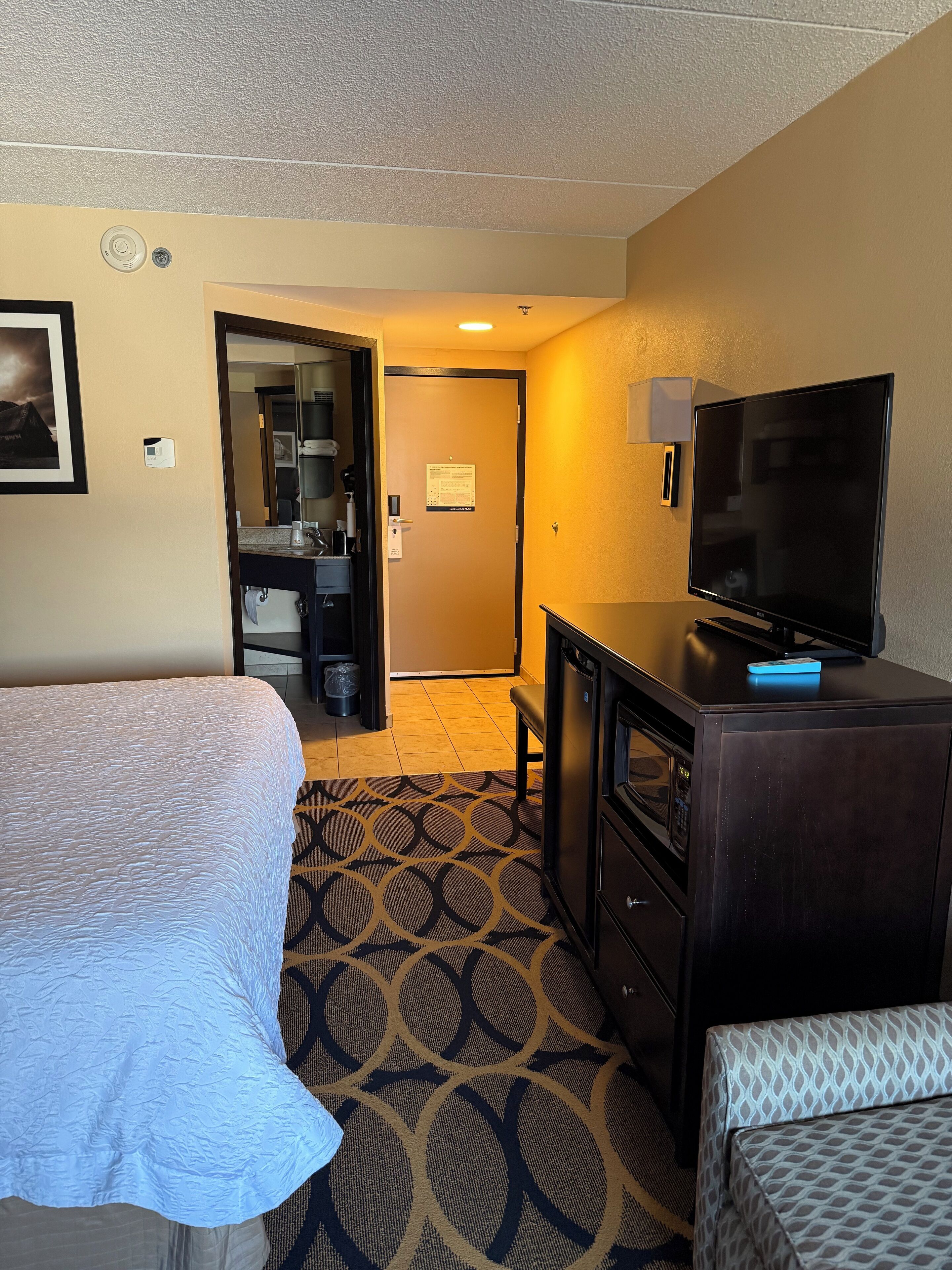 Habitación de hotel con cama, escritorio con televisor, microondas y baño visible a través de una puerta abierta.
