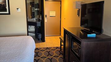 Habitación de hotel con cama, escritorio con televisor, microondas y baño visible a través de una puerta abierta.