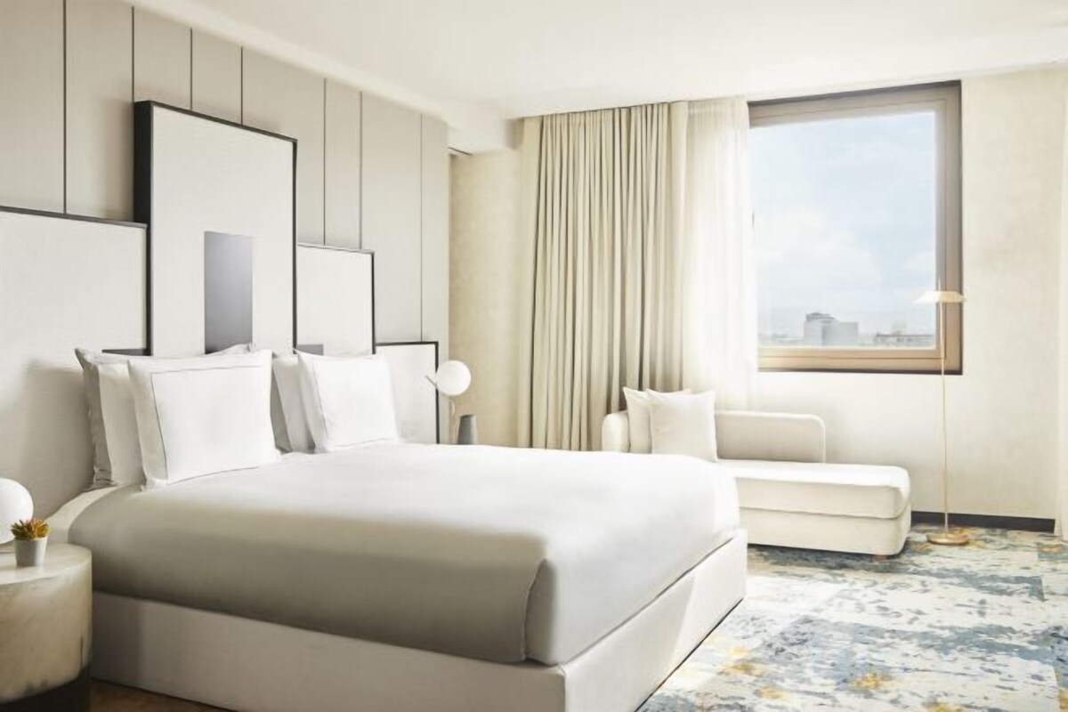 A hotel room with a large bed, a sofa, a window with curtains, and a city view.