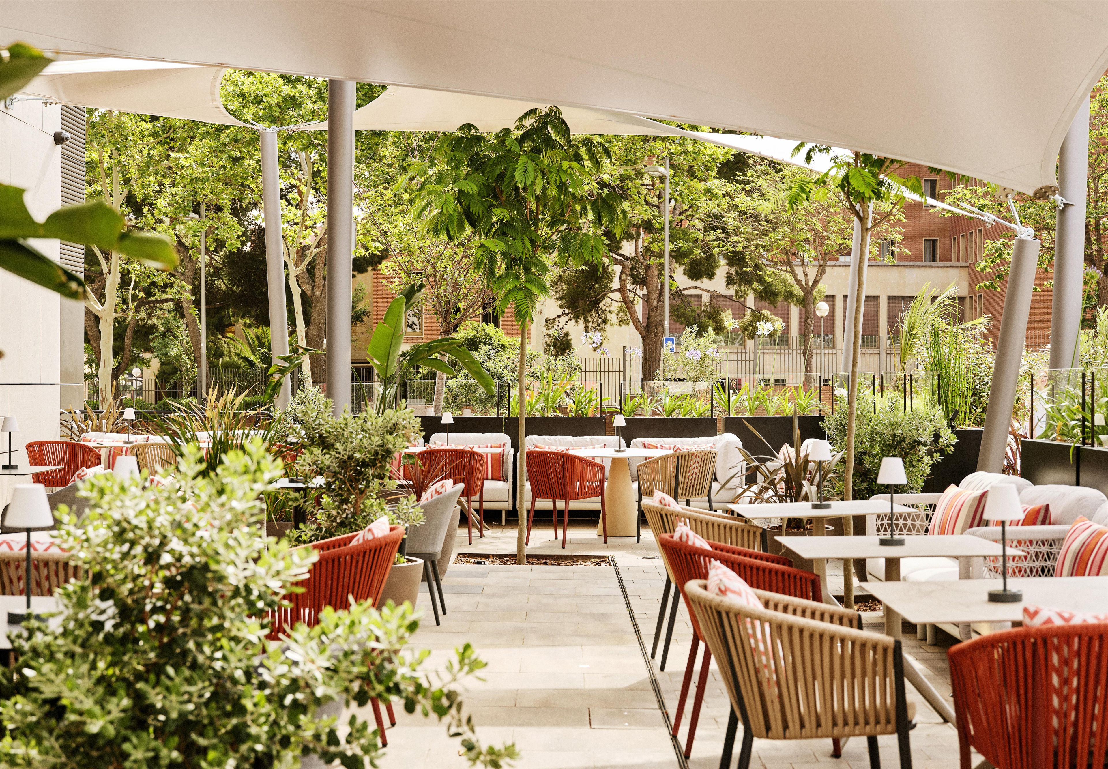An outdoor cafe area with tables and chairs, surrounded by greenery and a building in the background.