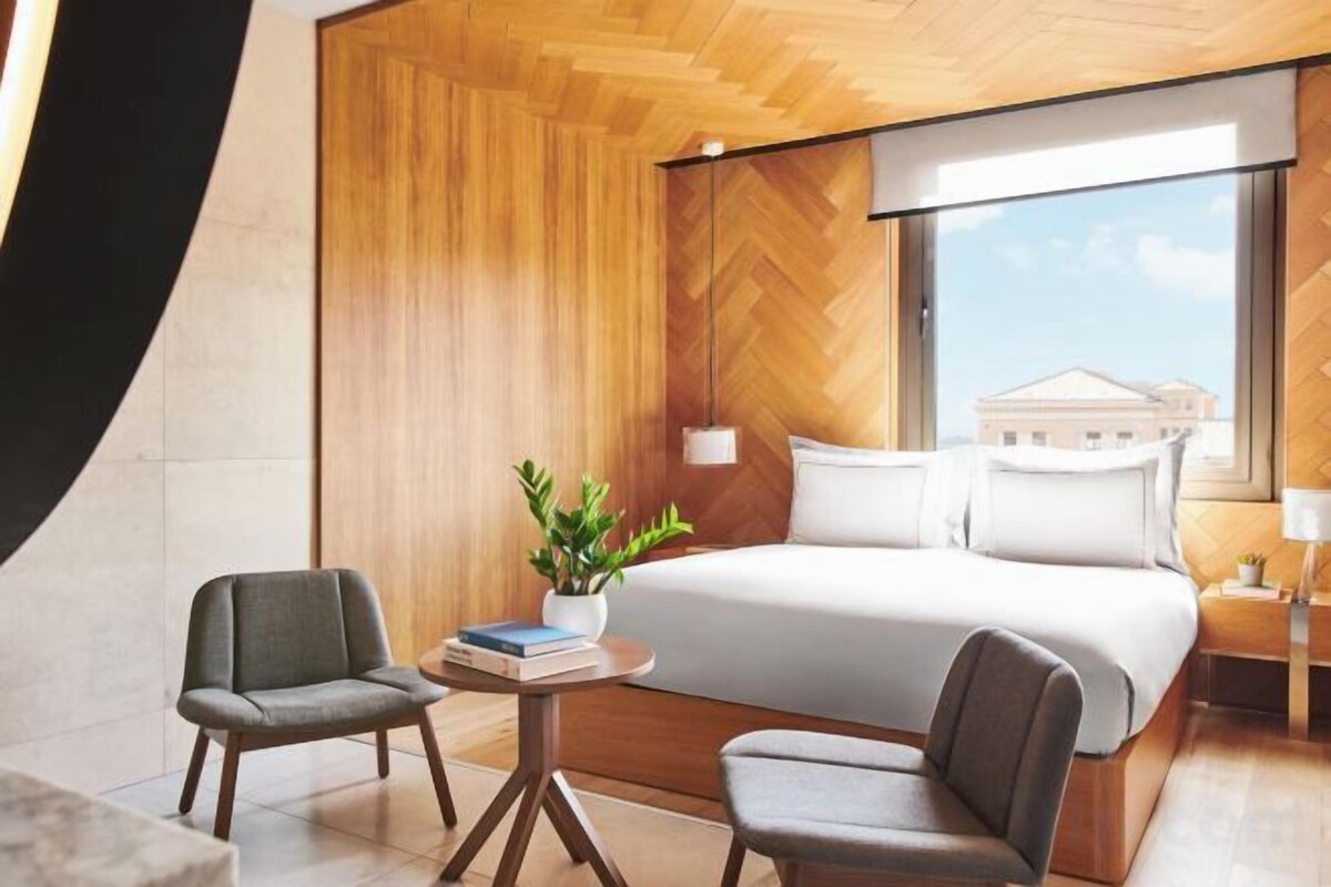 A modern hotel room with a large bed, two armchairs, a small table with a plant, and a view of a building through a large window.