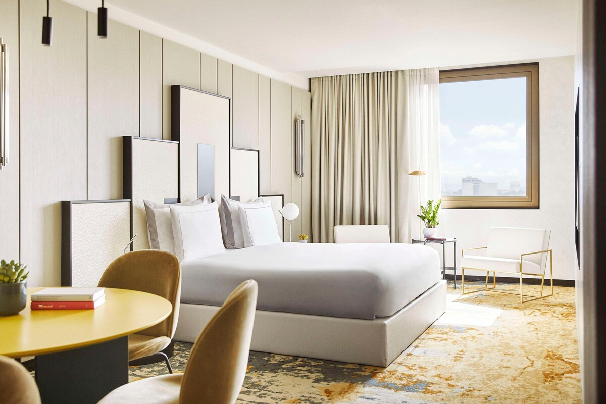 A modern hotel room with a large bed, a dining area with a table and chairs, a small side table with a plant, and a large window with a city view.