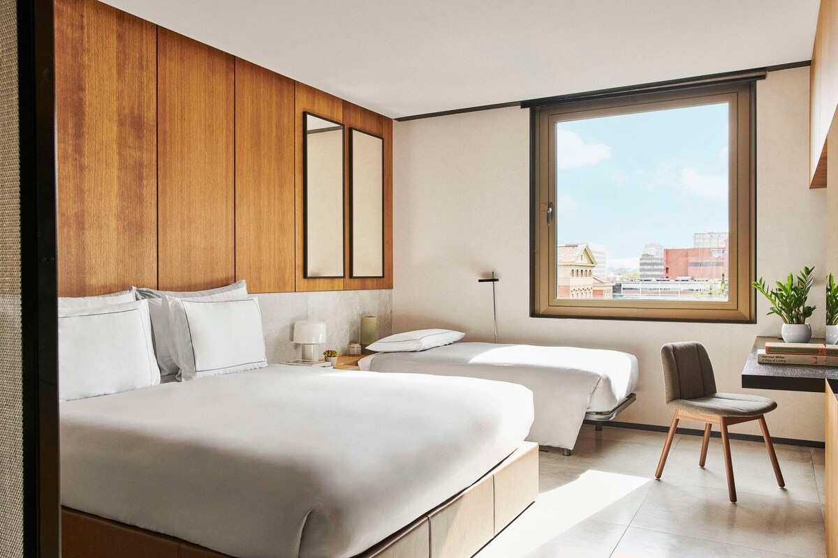 A modern hotel room with a large bed, a desk with a chair, a small table with a plant, and a window with a city view.