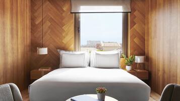 A modern hotel room with a large bed, two bedside tables, a window with a view, and a small table with a plant and books.