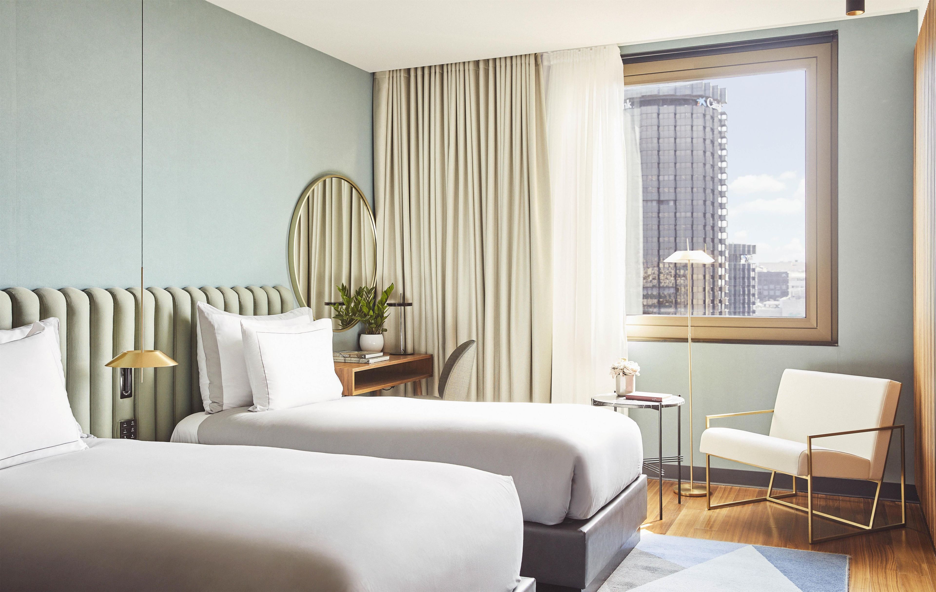 A hotel room with two beds, a desk, a chair, and a large window with a city view.