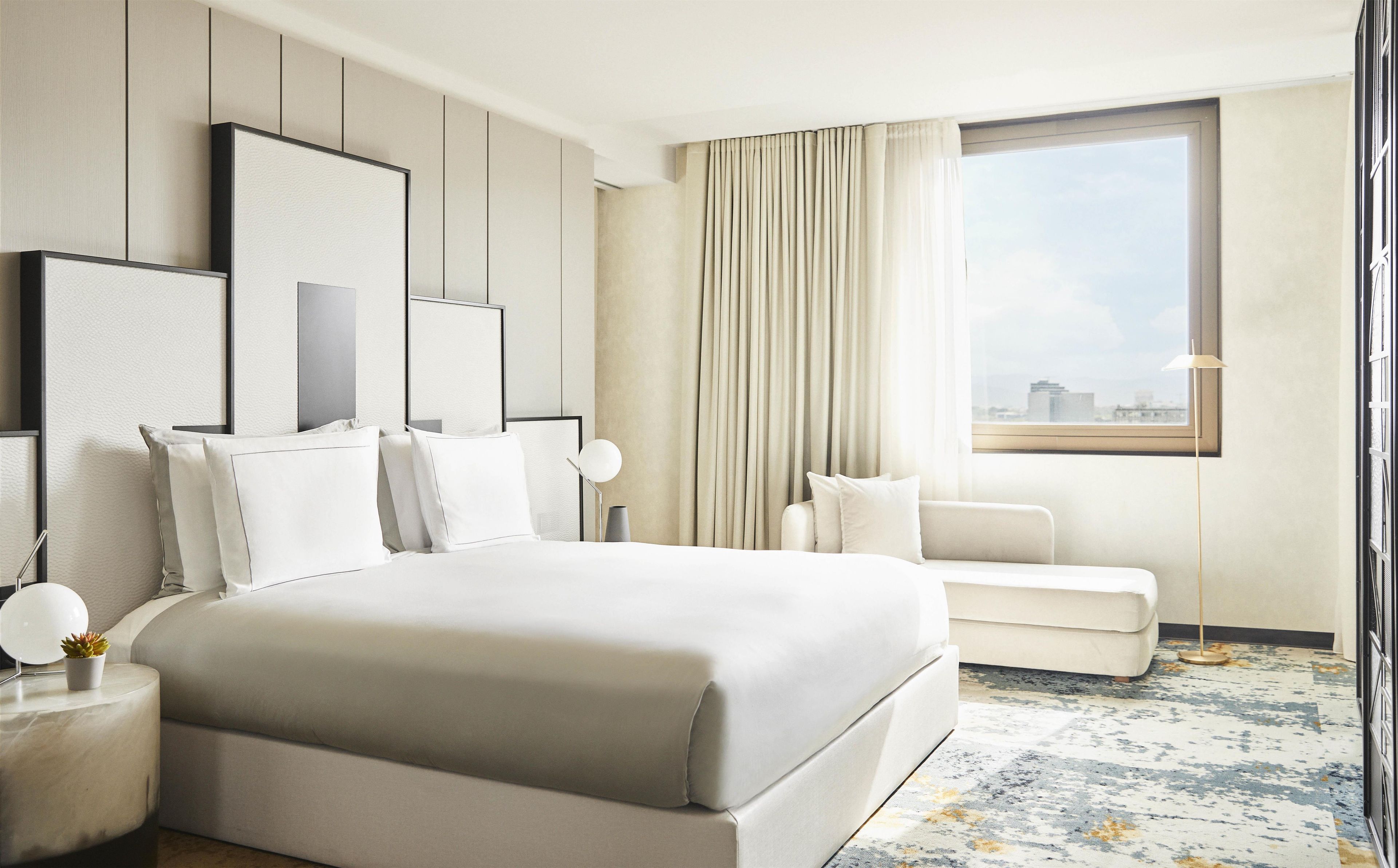 A hotel room with a large bed, a seating area, a window with a city view, and a modern design.