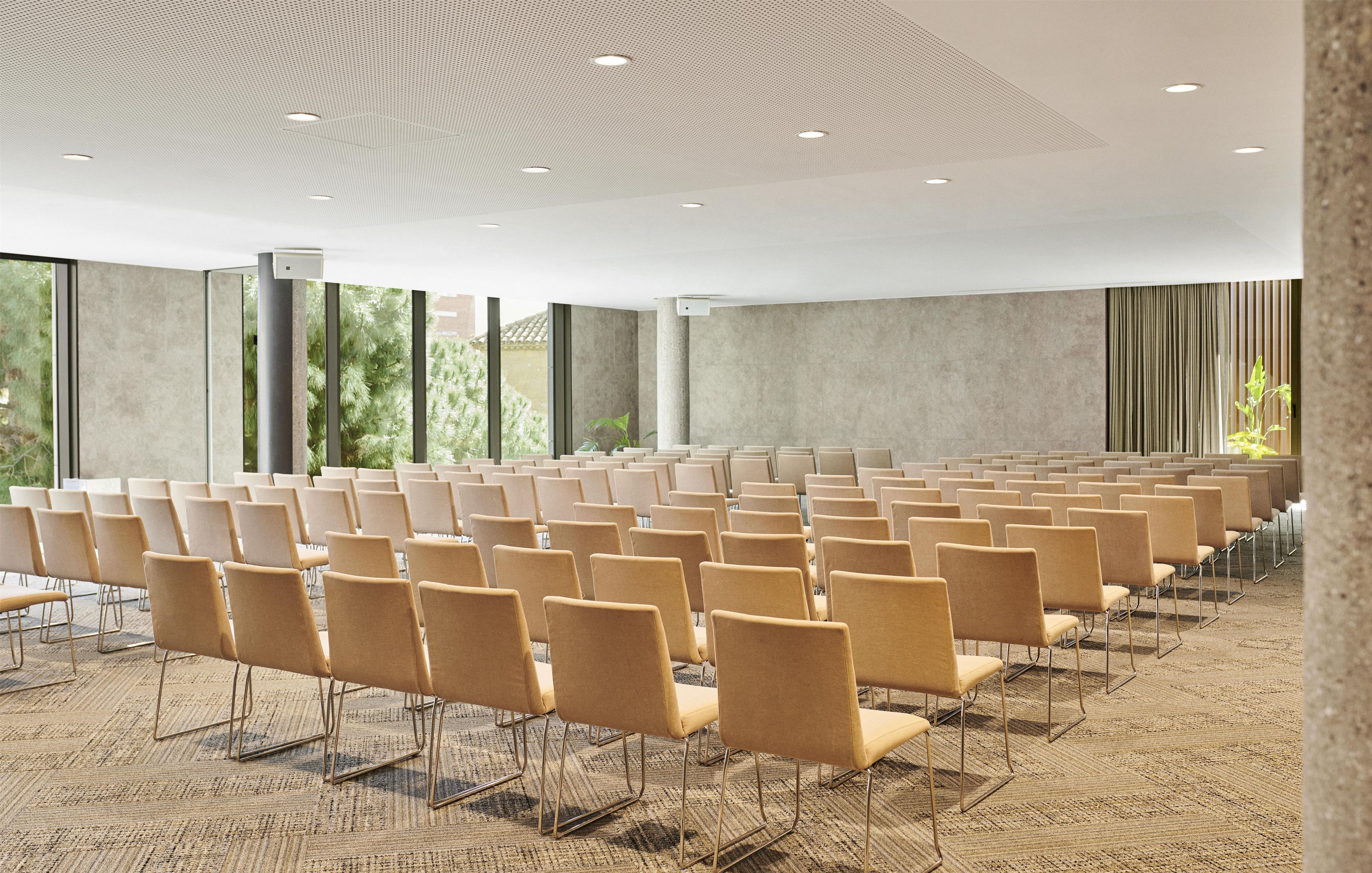 Un salón de conferencias con filas de sillas, ventanas amplias y un techo estampado.