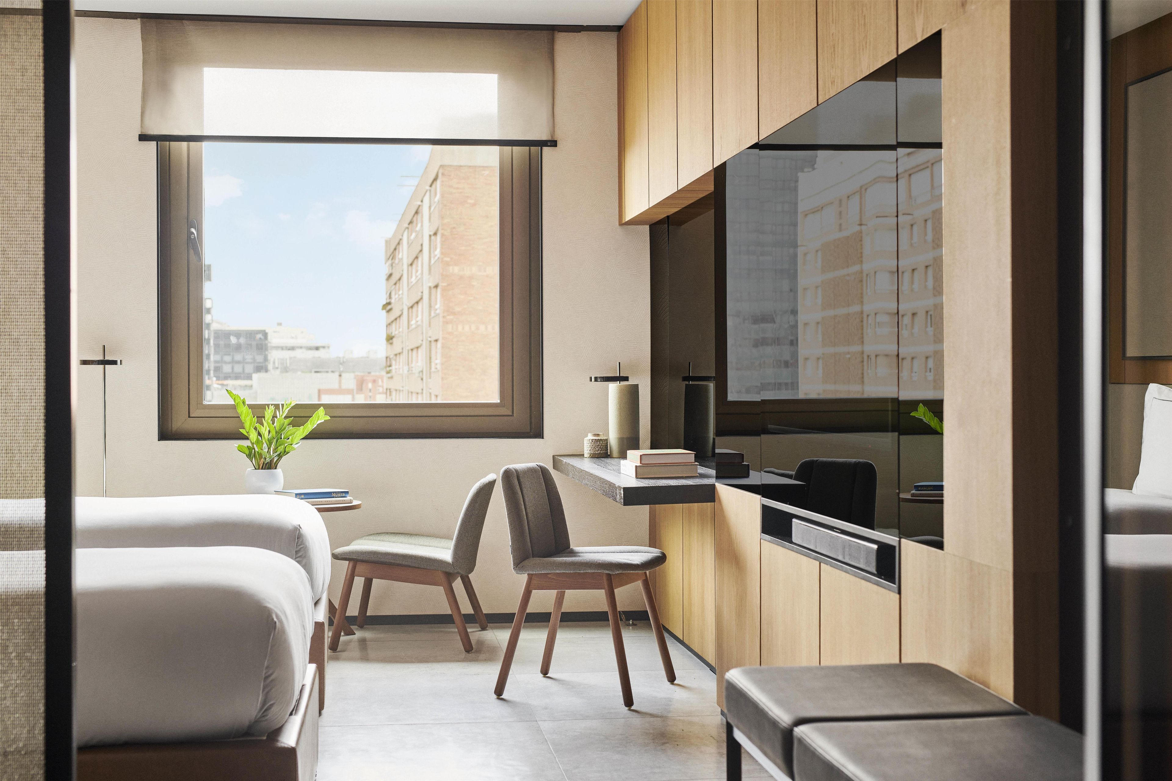 A modern hotel room with a large window, a desk, two chairs, a bed, and a small potted plant.