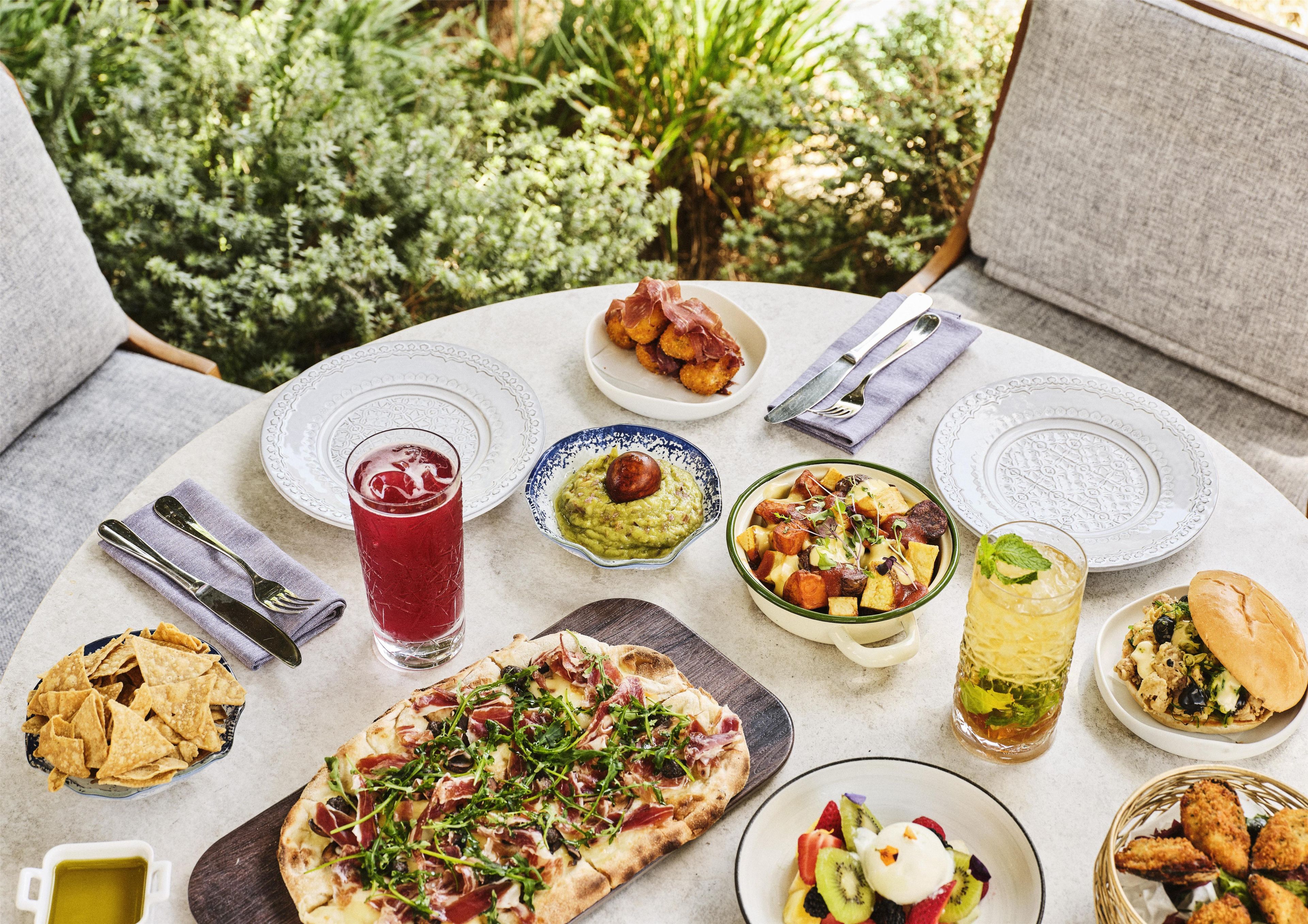 A table set outdoors with various dishes, including pizza, salad, and a drink.