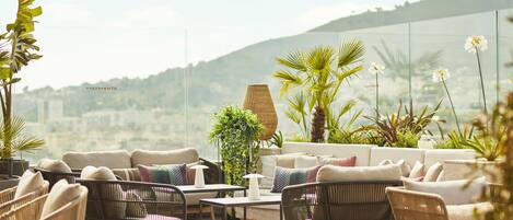 A rooftop terrace with wicker furniture, a table, and various plants.