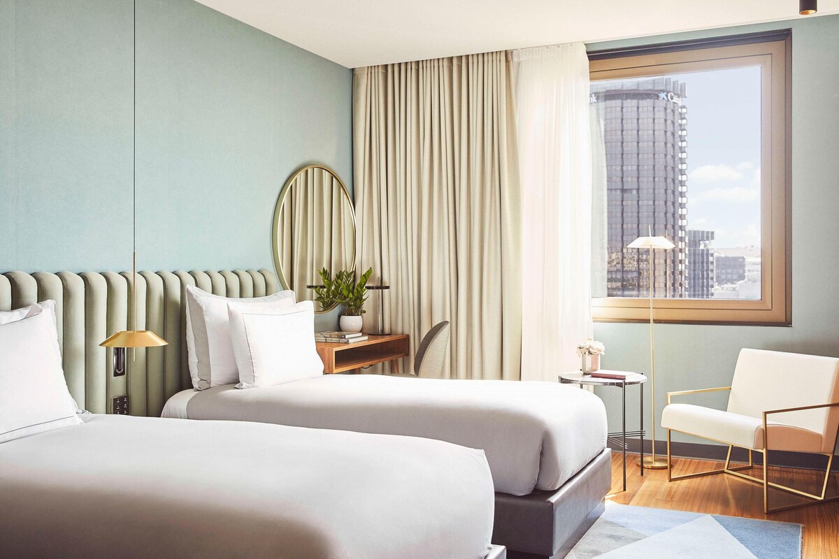 A hotel room with two beds, a desk, a chair, a plant, and a view of the city through a large window.