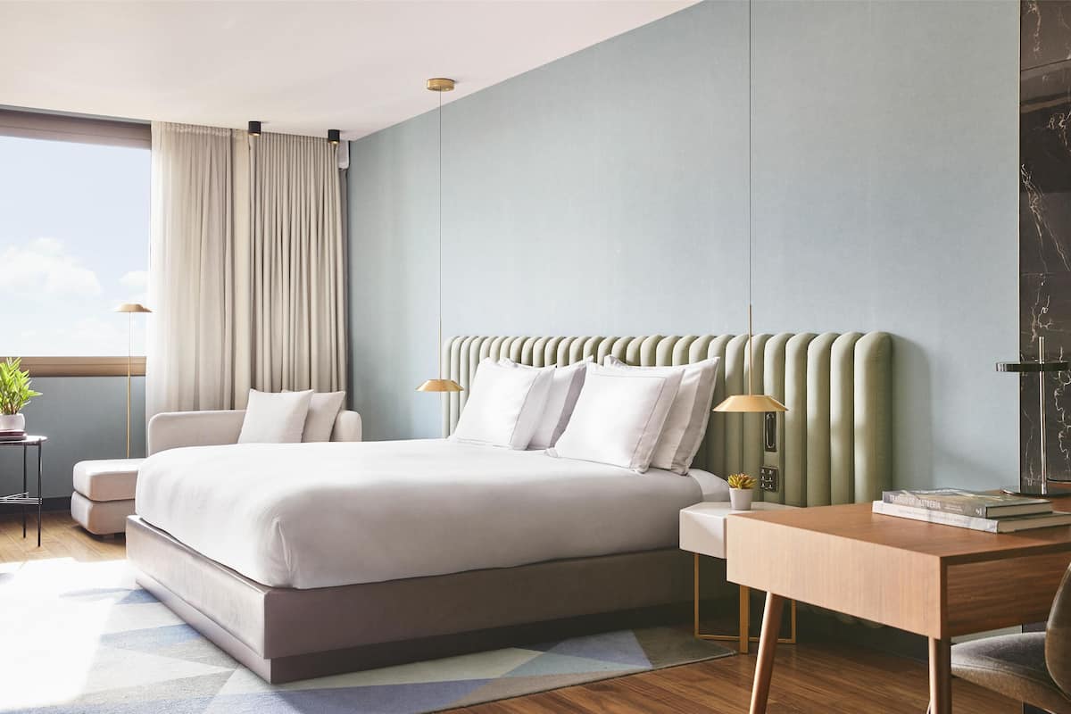 A modern bedroom with a large bed, a desk, and a chair. There is a window with curtains, a lamp, and a small table with a plant.