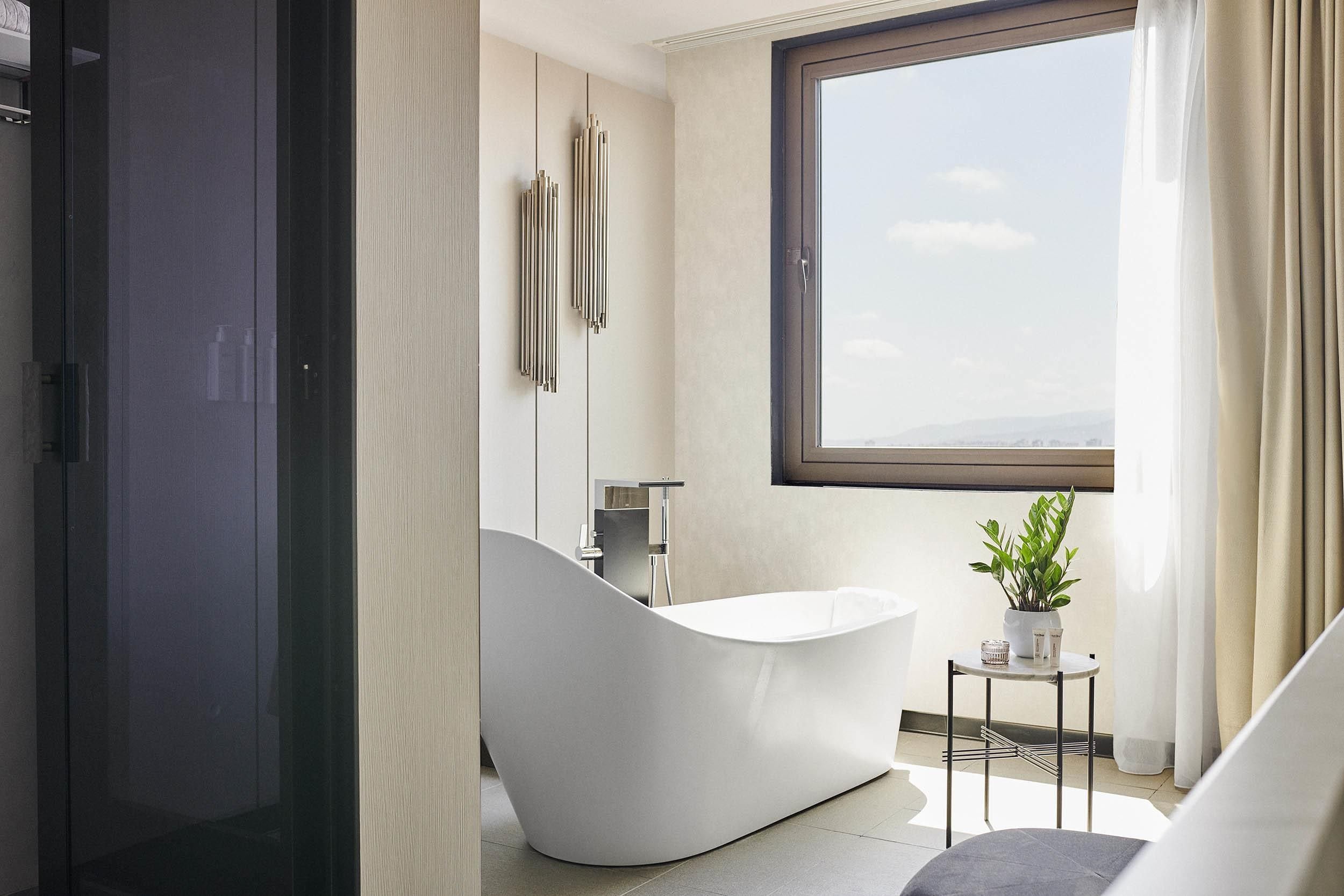 A modern bathroom with a freestanding bathtub, a window with a view, and a small table with a plant.