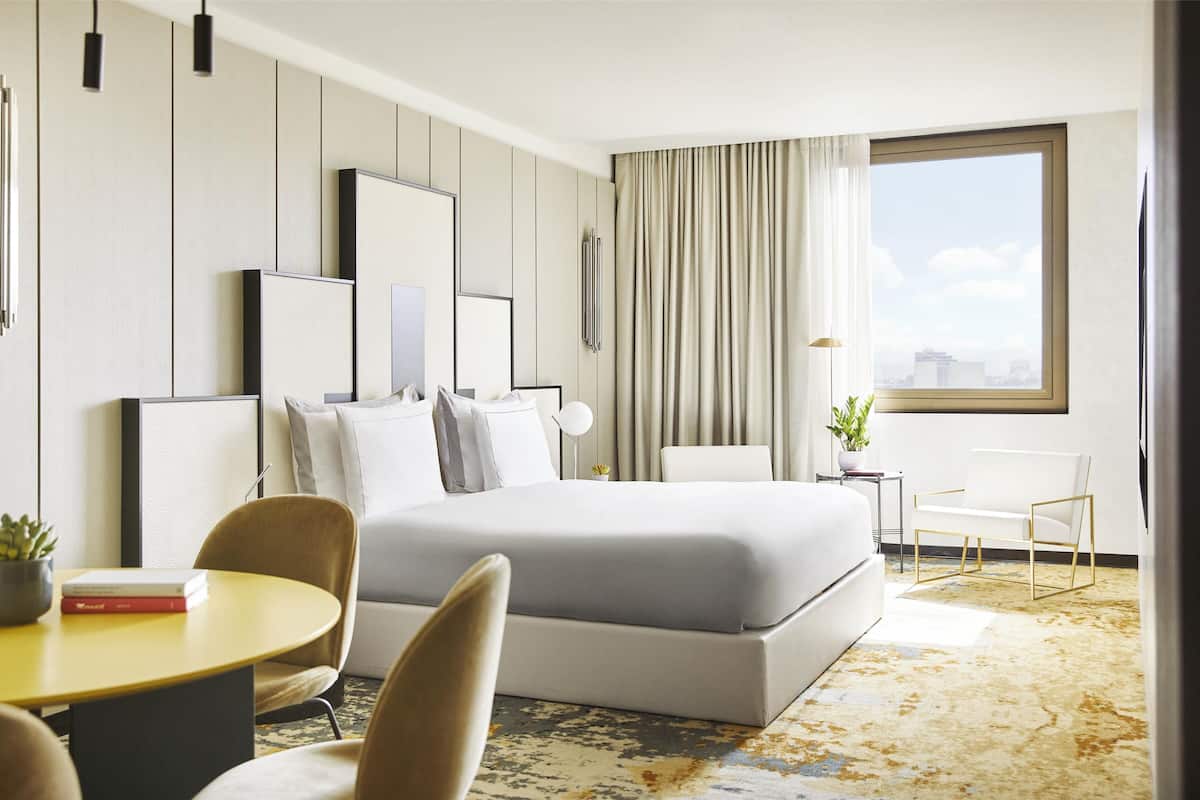 A modern hotel room with a large bed, a dining area, and a view of the city.