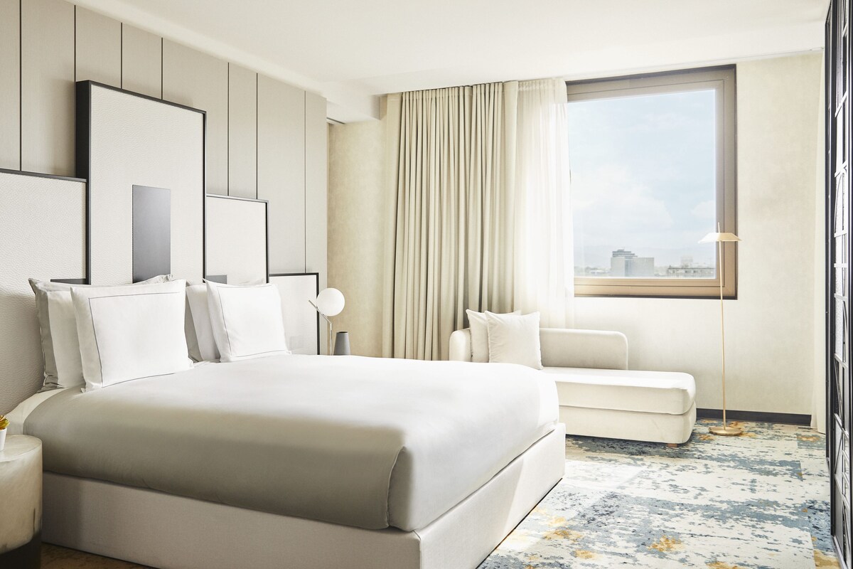 A modern hotel room with a large bed, a seating area, and a view of the city.