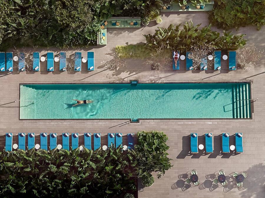 Vista aérea de una piscina rectangular con un solo nadador, rodeada de filas de sillas reclinables azules y una pequeña zona de estar con mesas y sillas.