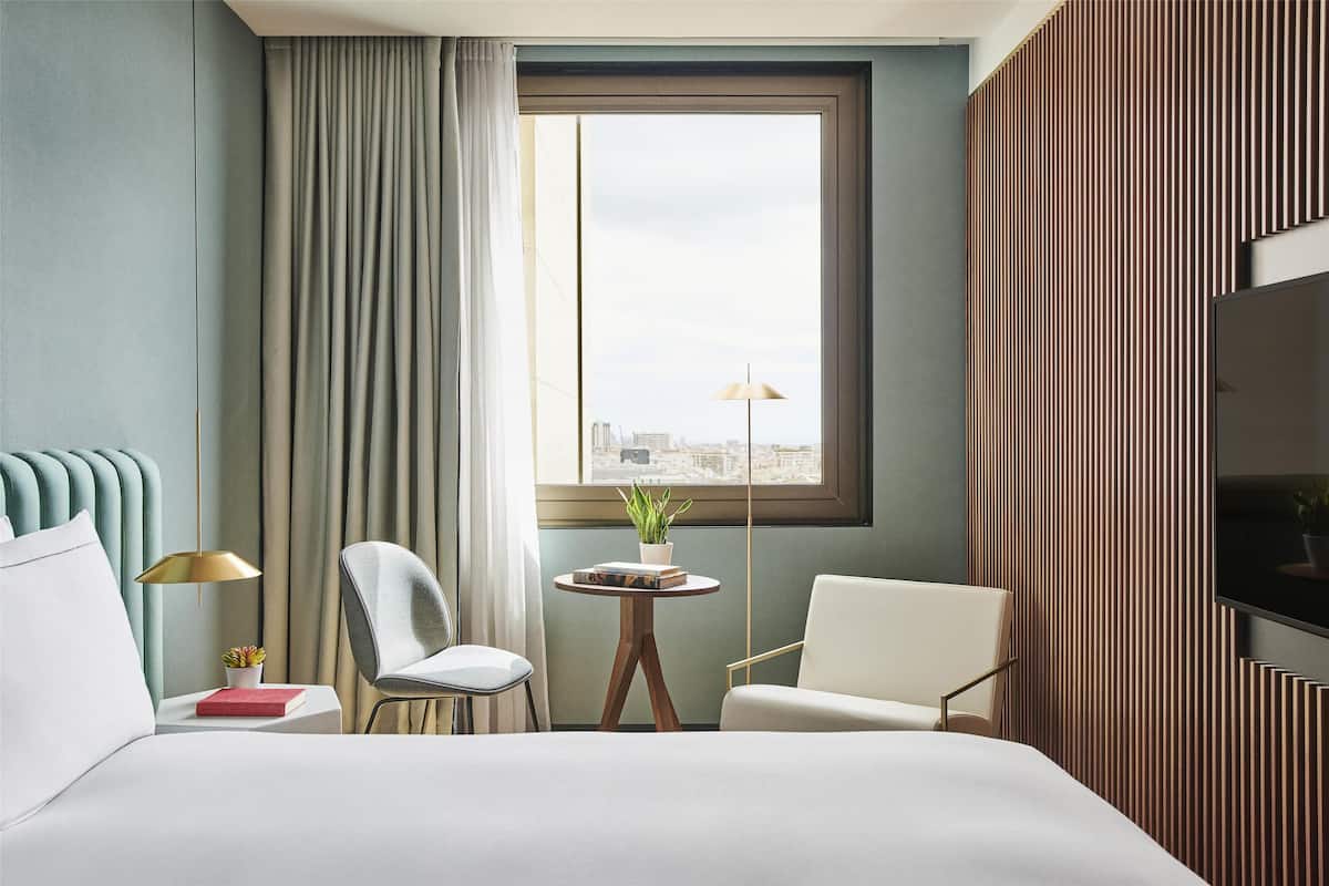 A hotel room with a bed, two chairs, a small table with a plant, a lamp, and a TV on the wall.