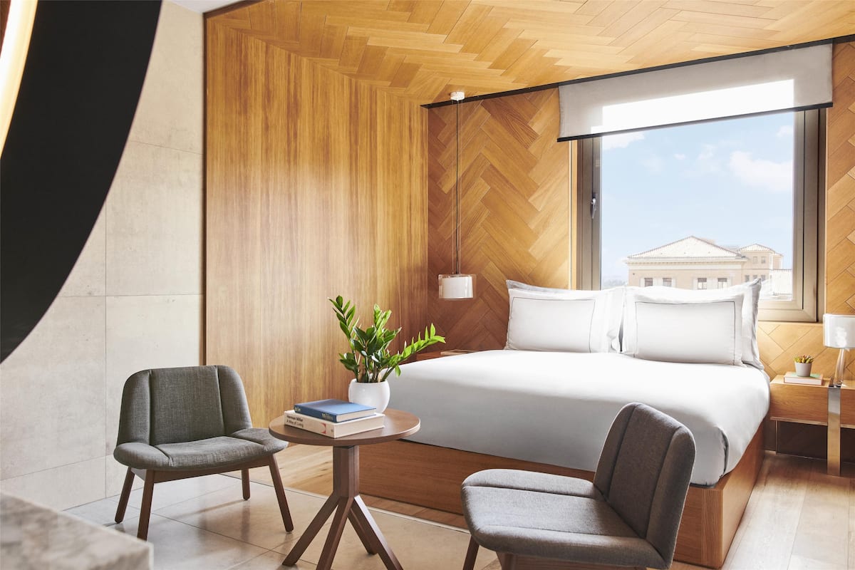 A modern hotel room with a large bed, two armchairs, a small table with a plant, and a view of a building through a large window.