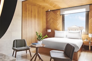 A modern hotel room with a large bed, two armchairs, a small table with a plant, and a view of a building through a large window.