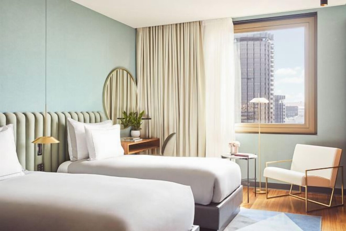 A hotel room with two beds, a desk, a chair, a window with a city view, and a large mirror.