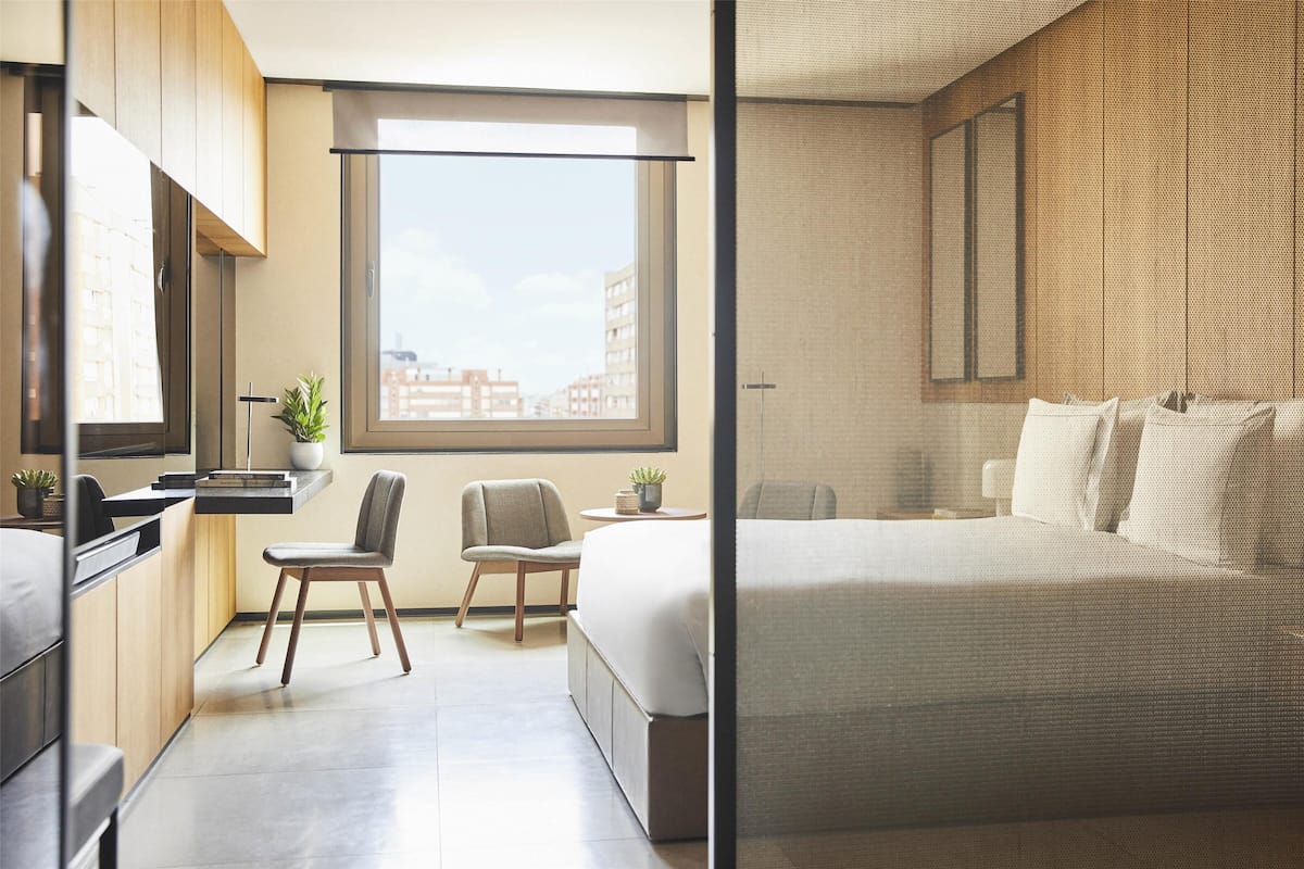 A modern hotel room with a large window, a bed, two chairs, a small table, and a potted plant.