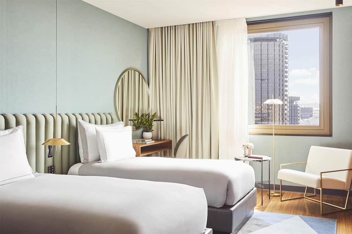 A modern hotel room with two beds, a desk, a chair, and a view of the city through large windows.