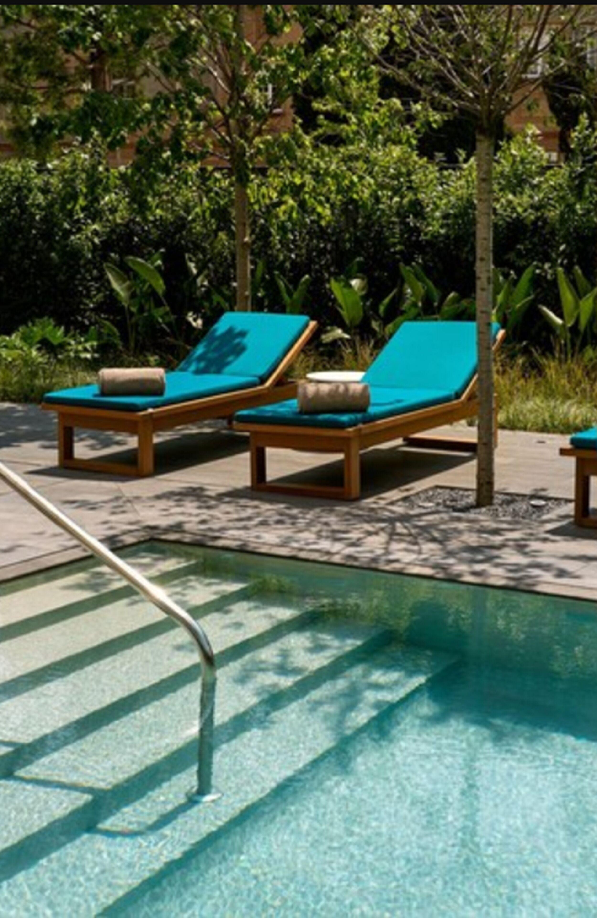 A poolside area with two wooden lounge chairs with blue cushions, surrounded by lush greenery.