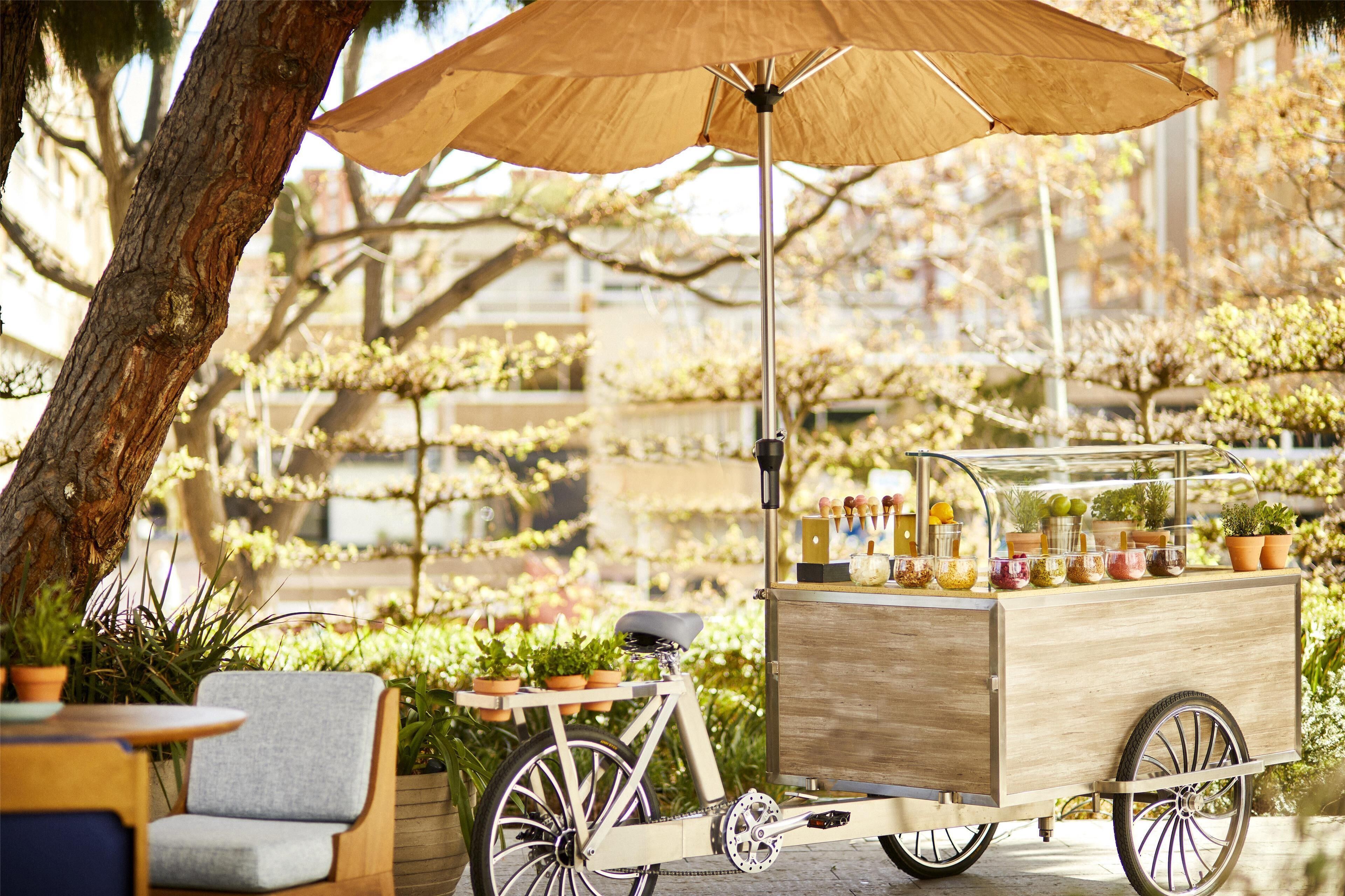 A stationary bicycle cart with a canopy, surrounded by potted plants and outdoor seating.