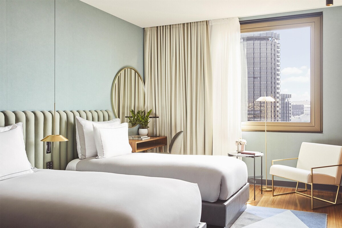 A hotel room with two beds, a desk, a chair, a potted plant, and a view of the city through a large window.